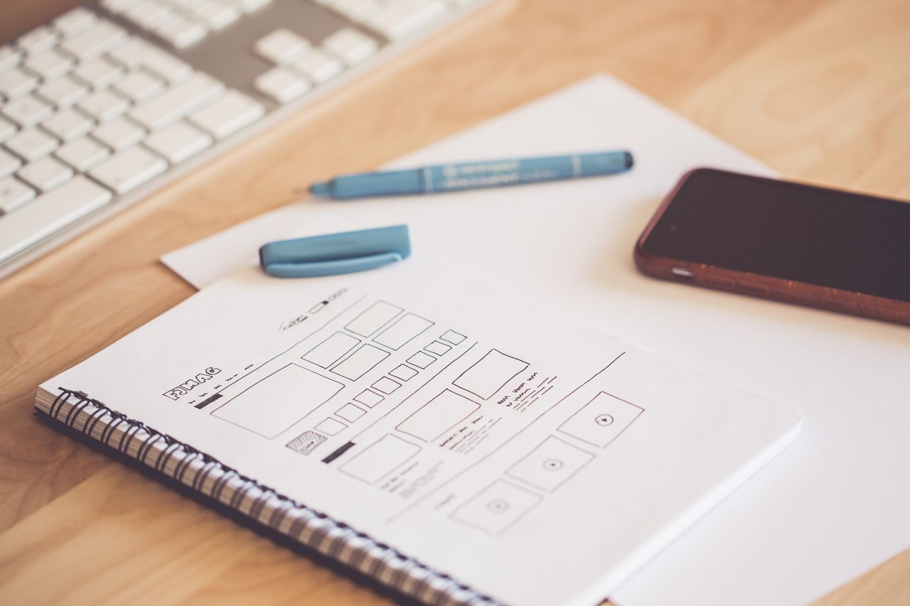 Mastering the First Impression: Your intriguing post title goes here A neat workspace featuring a sketchpad with wireframes, a smartphone, and a keyboard on a wooden desk.